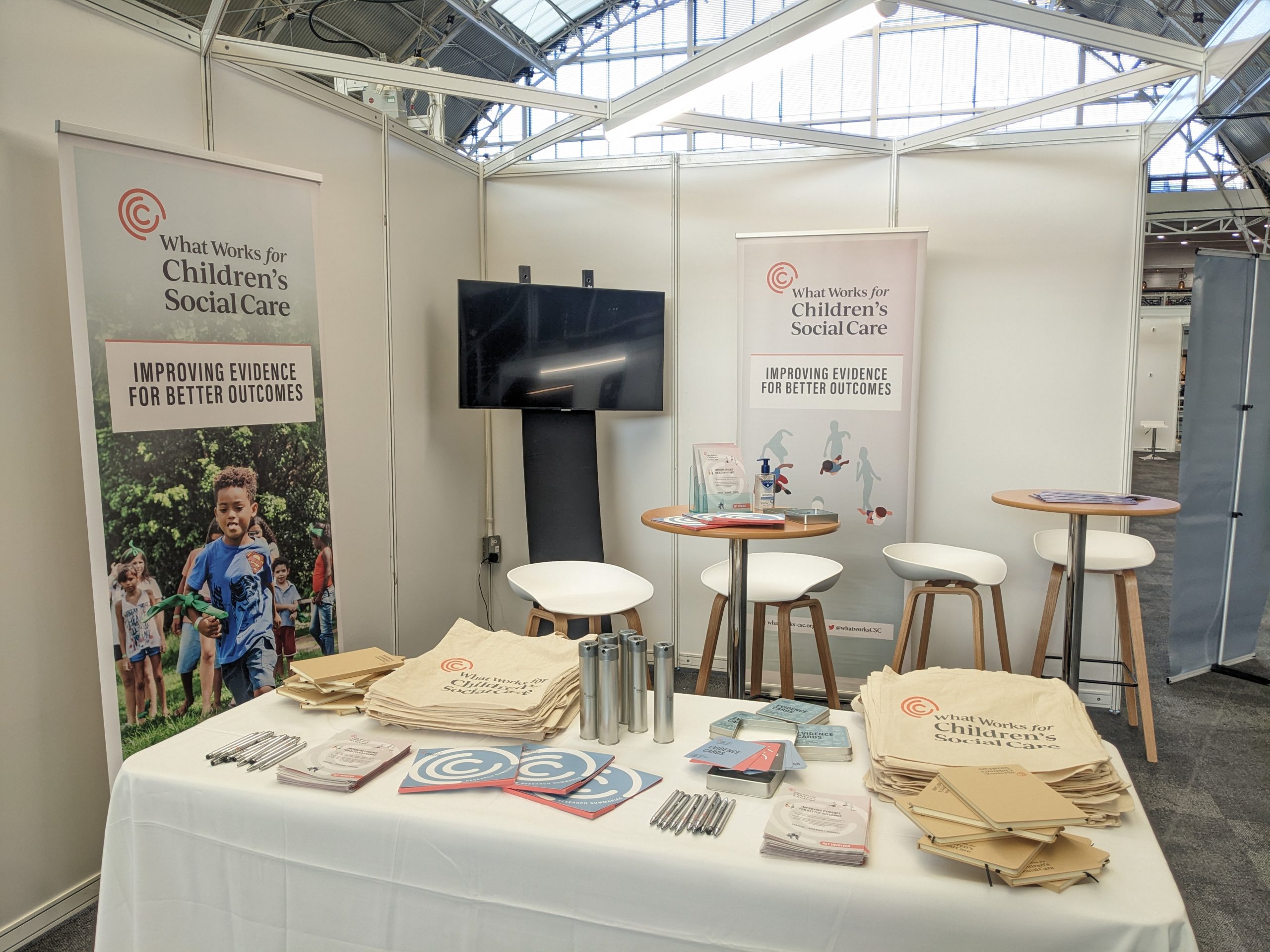 Our stand at Community Care live 2022. A long white table with piles of merchandise and leaflets on it, whilst banners with our branding on line the back of the stand