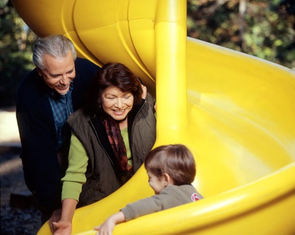 A child slides down a yellow slide watched by a smiling mature couple